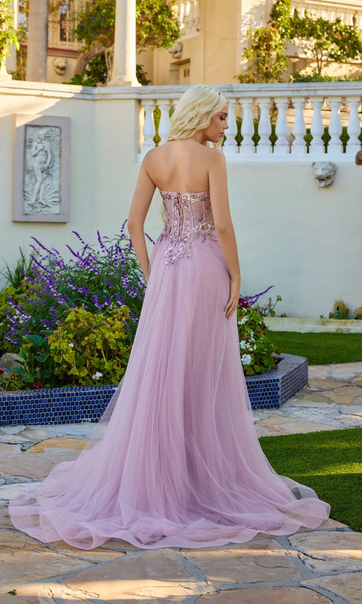 A woman stands outdoors on a stone path, wearing the Juliet JT2640G long prom dress—strapless, floor-length light purple tulle with floral embellishments, a sheer skirt, and lace-up back—facing away from the camera.